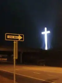 Night photograph of a yellow ONE WAY street sign pointing toward a large glowing white cross in the distance